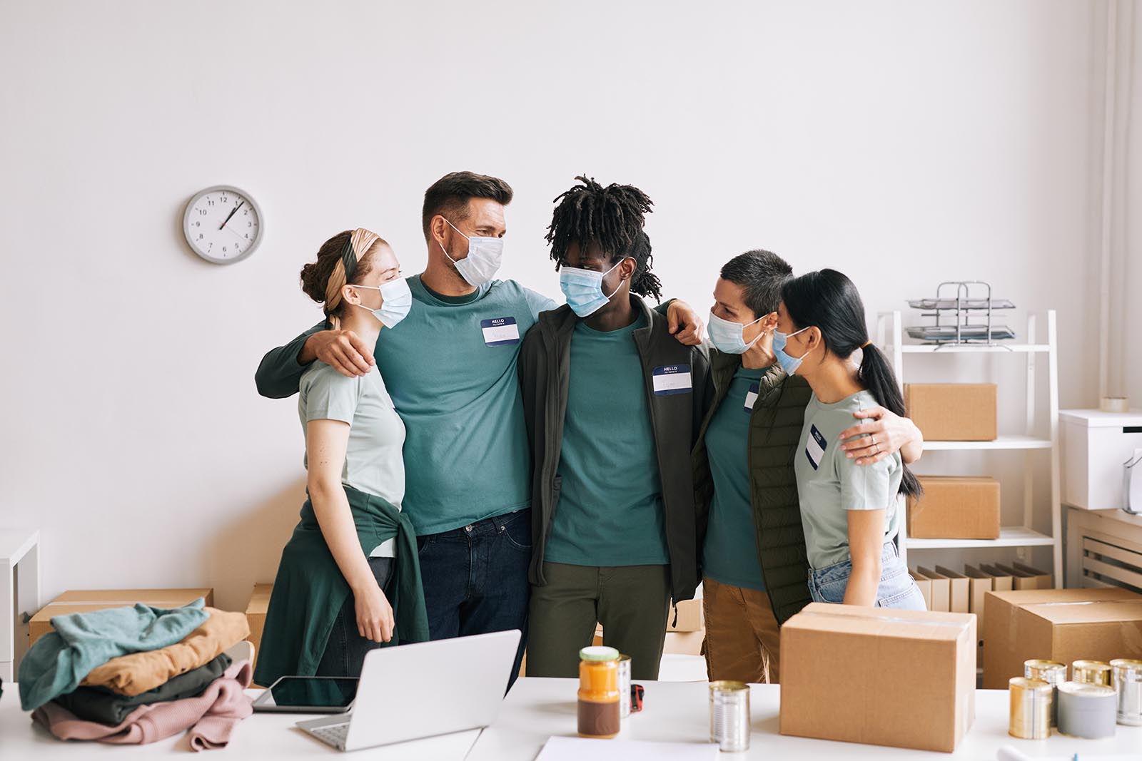 Diverse team of volunteers wearing masks while embracing at help and donation event, copy space